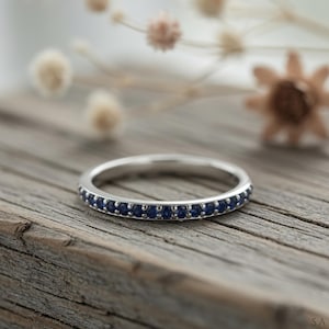 May include: A silver ring featuring a band of small, round, dark blue gemstones. The ring is set on a weathered wooden surface, with dried flowers in the background. A delicate piece of jewellery.