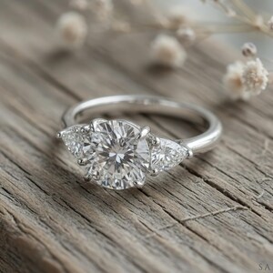 May include: A silver ring with a large, round, clear gemstone flanked by two smaller, pear-shaped stones. The ring sits on a weathered wooden surface, with delicate white flowers in the background. The ring is a classic design.