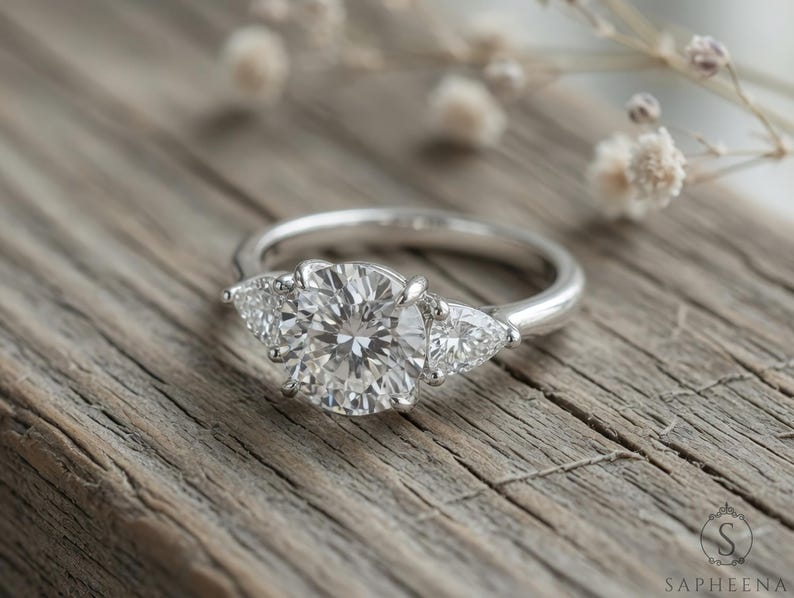 May include: A silver ring with a large, round, clear gemstone flanked by two smaller, pear-shaped stones. The ring sits on a weathered wooden surface, with delicate white flowers in the background. The ring is a classic design.