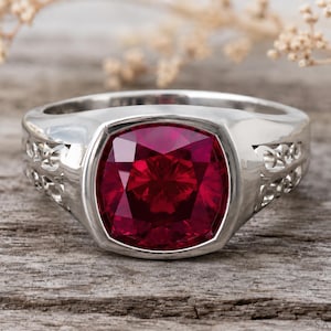 May include: A silver-toned ring featuring a large, cushion-cut, deep red gemstone set in a smooth bezel. The band displays intricate, openwork filigree detailing along the sides, providing a decorative contrast to the polished metal finish.