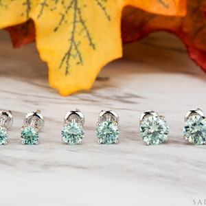 May include: Six pairs of round, light blue gemstone stud earrings in varying sizes are displayed on a marble surface. The earrings have silver posts and settings. A yellow and orange leaf is in the background. The brand name "SAPHEENA" is in the bottom right corner.