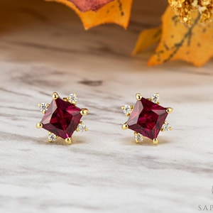 May include: A pair of gold stud earrings. Each earring has a square, deep red gemstone at the centre, encircled by smaller, clear gemstones and gold accents. The earrings are displayed on a marble-effect surface.
