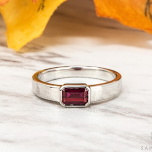 May include: A silver ring featuring a rectangular, deep red gemstone. The ring has a brushed metal finish and a simple design. The gemstone is set in a bezel, and the ring is displayed on a white surface.