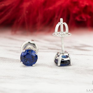May include: A pair of silver stud earrings featuring a round blue sapphire gemstone. The earrings have a screw-back fastening.