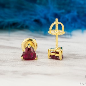 May include: A pair of gold heart-shaped stud earrings with red ruby gemstones. The earrings have a post and butterfly back closure.