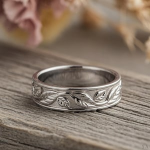 May include: A silver ring with a detailed leaf pattern. The ring is set against a rustic wooden surface, with soft, blurred floral elements in the background. The ring's design features a repeating pattern of leaves and vines.