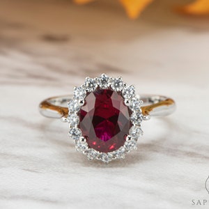 May include: A silver ring with a large, oval-shaped ruby gemstone surrounded by a halo of smaller, clear gemstones.