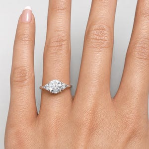 May include: A close-up of a hand wearing a silver ring with a large, round, clear center stone and two smaller stones on either side. The ring is set on a light skin tone.