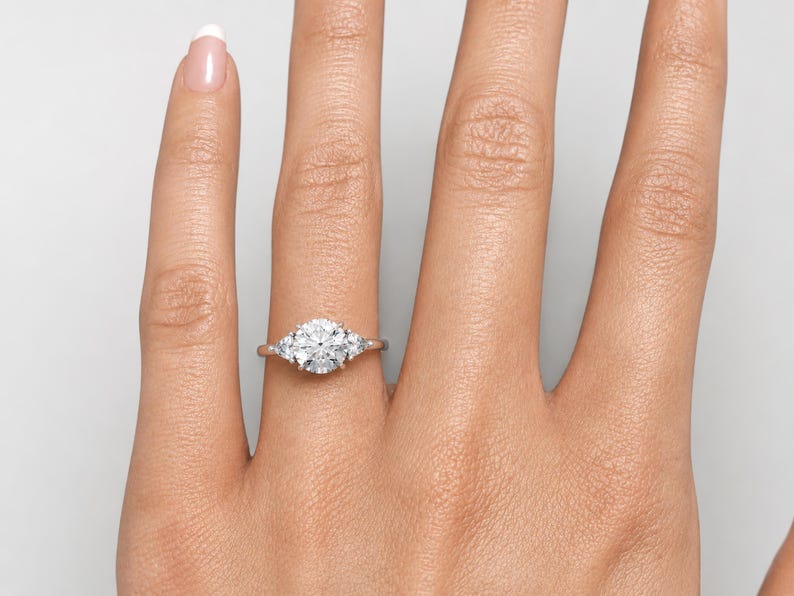 May include: A close-up of a hand wearing a silver ring with a large, round, clear center stone and two smaller stones on either side. The ring is set on a light skin tone.