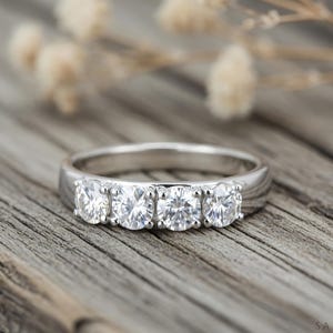 Può includere: Un anello d'argento con cinque gemme rotonde e trasparenti disposte in fila. L'anello è presentato su una superficie di legno invecchiato, con elementi floreali sfocati sullo sfondo. È un gioiello.