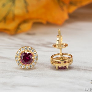 May include: A pair of gold earrings featuring a red gemstone set in a halo of smaller white gemstones.