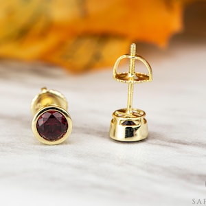Könnte beinhalten: Ein Paar goldene Ohrstecker mit einem roten Edelstein in der Mitte. Die Ohrringe haben einen Stift und einen Schmetterlingsverschluss.