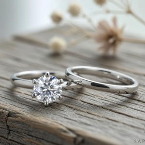 Puede incluir: Dos anillos de plata sobre una superficie de madera. Uno presenta una gran gema redonda y transparente con seis garras. El otro es una banda lisa y pulida. Flores secas en el fondo.