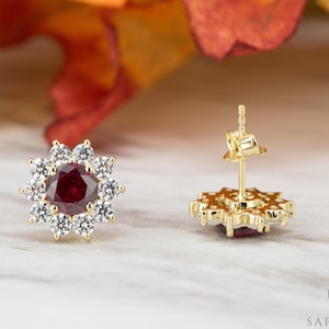 Peut inclure: Une paire de boucles d'oreilles en or avec une pierre précieuse rouge entourée de pierres de zircone cubique transparentes. Les boucles d'oreilles ont une fermeture à tige.