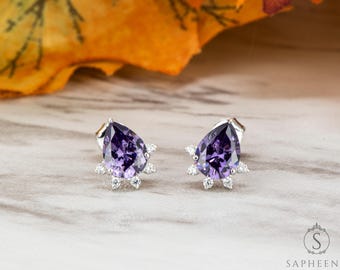 May include: A pair of silver stud earrings featuring pear-shaped, deep purple gemstones. Each earring is accented with small, clear crystals around the base of the purple stone. The earrings are set against a neutral background.