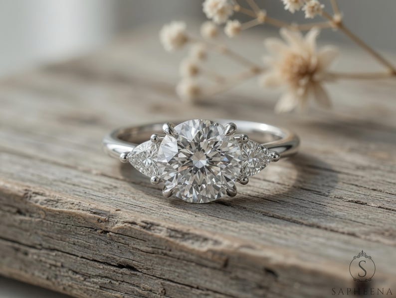 May include: A silver engagement ring with a large, round, clear center stone flanked by two smaller, pear-shaped stones. The ring is set on a rustic wooden surface, with dried flowers in the background.