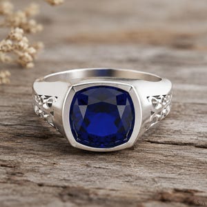 Peut inclure: Une bague argentée ornée d'une grande pierre précieuse bleu profond taille coussin, sertie dans une monture lisse. L'anneau présente des détails en filigrane ajouré sur les côtés, offrant un contraste décoratif avec le métal poli.