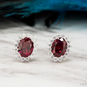 May include: A pair of silver earrings featuring a large oval ruby gemstone set within a halo of smaller clear gemstones.