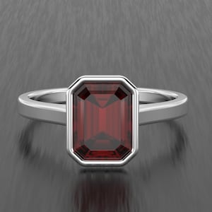May include: A silver ring with a rectangular, deep red gemstone. The gemstone is set in a silver bezel, and the band is plain, polished silver. The ring is shown against a dark, textured background.