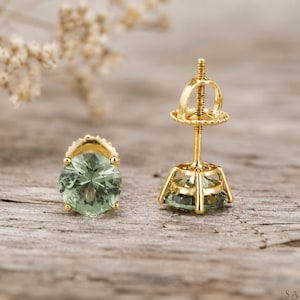 May include: A pair of stud earrings featuring round-cut, light green gemstones set in yellow gold. One earring is displayed from the front, while the other is shown from the side, revealing the gold post and the threaded screw-back mechanism.