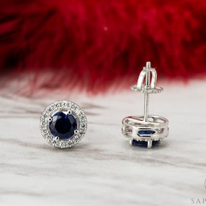May include: A pair of silver stud earrings featuring a blue sapphire centre stone surrounded by a halo of smaller diamonds.