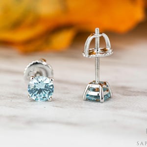 May include: A pair of silver stud earrings with a light blue round gemstone in each. The earrings have a post and butterfly back.