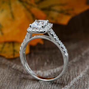 以下が含まれることがあります： 正方形のセンター石と小さな石のハローが付いたシルバーの婚約指輪のクローズアップ。バンドにも小さな石が飾られています。リングは、秋の葉のぼやけた背景に設定されています。