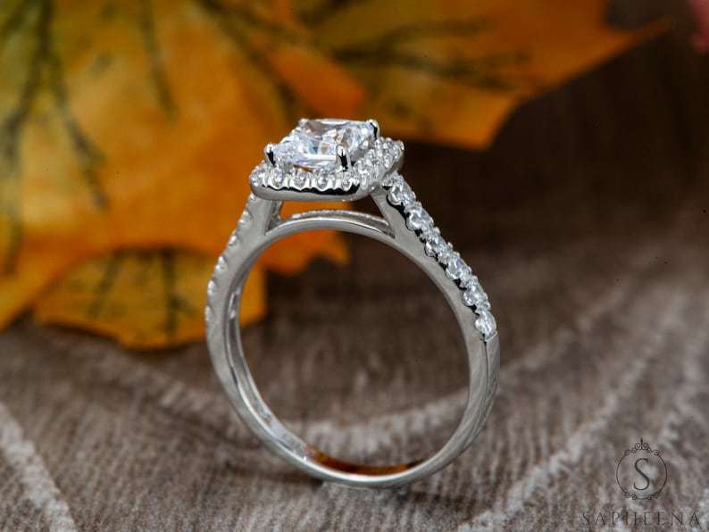 以下が含まれることがあります： 正方形のセンター石と小さな石のハローが付いたシルバーの婚約指輪のクローズアップ。バンドにも小さな石が飾られています。リングは、秋の葉のぼやけた背景に設定されています。