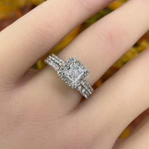 以下が含まれることがあります： 正方形のセンター石と小さな石のハローが付いたシルバーの婚約指輪セット。お揃いのバンドにも小さな石が付いています。リングは明るい肌の色合いで表示されています。