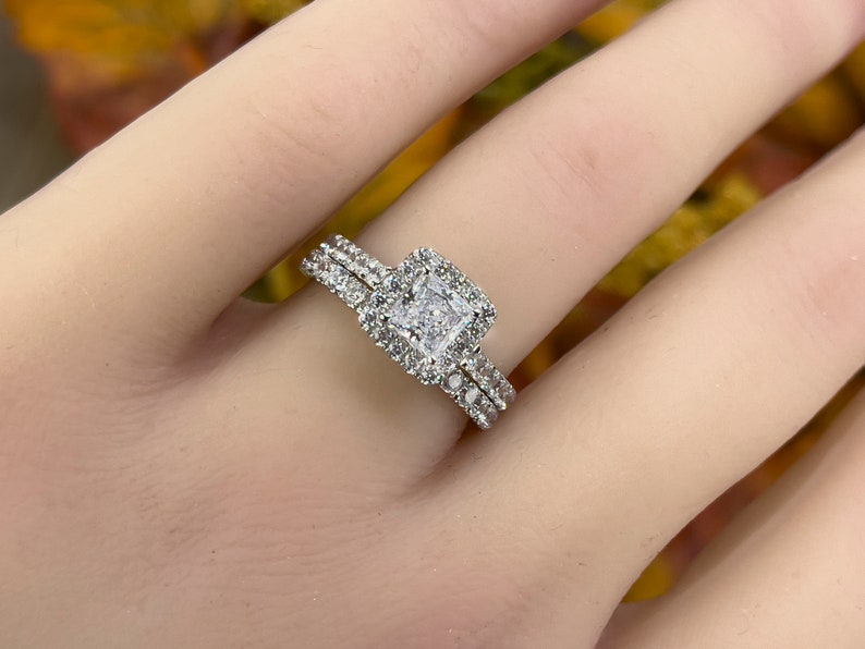以下が含まれることがあります： 正方形のセンター石と小さな石のハローが付いたシルバーの婚約指輪セット。お揃いのバンドにも小さな石が付いています。リングは明るい肌の色合いで表示されています。