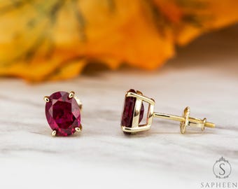 May include: A pair of gold stud earrings featuring oval-cut, deep red gemstones. The earrings have a four-prong setting and a screw-back closure. The background is blurred, with hints of yellow and orange.