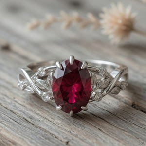 May include: A silver ring featuring a large, oval-cut, deep red gemstone. The band has an intricate leaf design, embellished with small, clear gemstones. The ring is displayed on a rustic wooden surface.