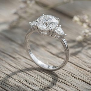 May include: A silver engagement ring with a large, round center stone and two smaller, triangular side stones. The ring is set on a light-colored wooden surface, with soft lighting and a blurred background.