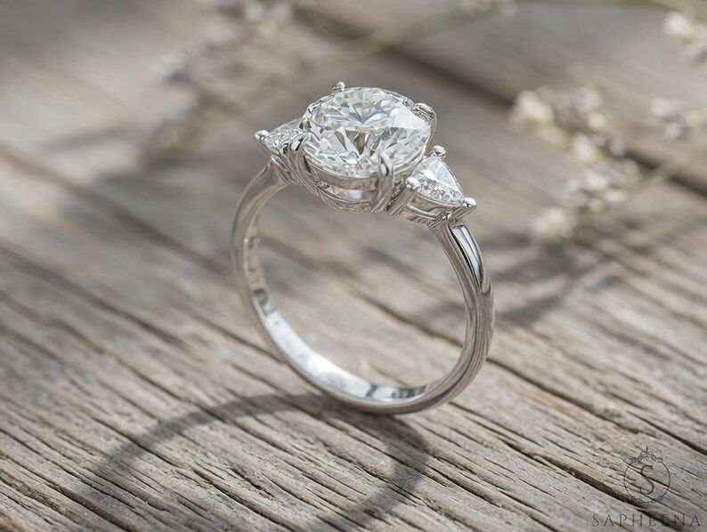 May include: A silver engagement ring with a large, round center stone and two smaller, triangular side stones. The ring is set on a light-colored wooden surface, with soft lighting and a blurred background.