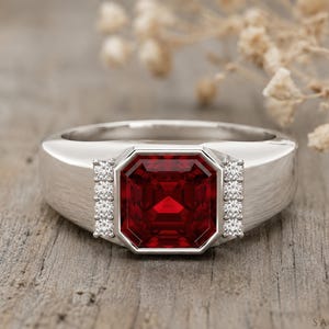 May include: A silver-toned ring featuring a large, square-cut, deep red gemstone at the centre. The stone is flanked on both sides by a vertical row of four small, clear, round-cut stones set into the band. The band has a smooth, polished finish.