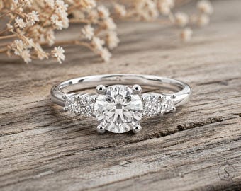 May include: A silver engagement ring with a large, round, clear diamond center stone and two smaller diamond clusters on either side. The ring is set on a wooden surface with dried flowers in the background.