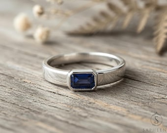May include: A silver ring with a rectangular, deep blue gemstone set in the center. The ring is on a weathered wooden surface, with soft, blurred background elements. The ring's design is simple and elegant, suitable for various occasions.