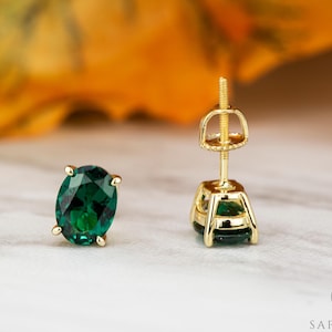 May include: A pair of gold-colored stud earrings with oval-cut, deep green gemstones. The earrings are displayed on a light-colored surface, with a blurred background of yellow and orange hues.