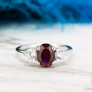 May include: A silver ring featuring a large oval ruby gemstone flanked by two smaller round diamonds.