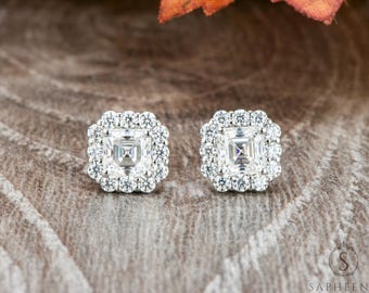 May include: A pair of square-cut diamond stud earrings. Each earring features a central square diamond surrounded by a halo of smaller, round diamonds. The earrings are set on a wooden surface.
