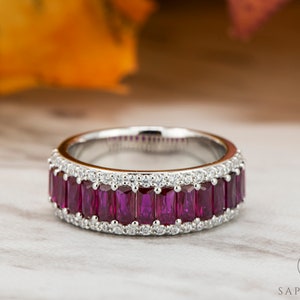 Può includere: Un anello a fascia d'argento con una fila di pietre di rubino rettangolari incastonate in una montatura a canale. L'anello è circondato da una fila di diamanti rotondi più piccoli.