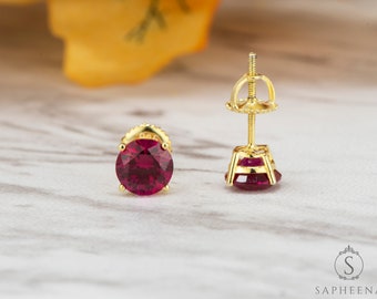 May include: A pair of gold stud earrings with a round, red gemstone. The earrings have a screw-back fastening.