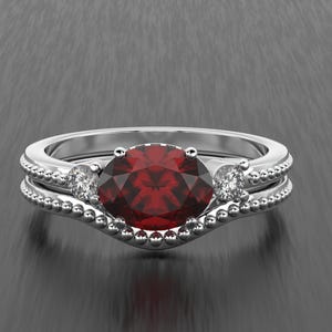 Puede incluir: Un anillo de plata con una gran gema roja oscura redonda y facetada, flanqueada por dos gemas claras más pequeñas. La banda presenta un diseño de cuentas. Debajo hay una banda curva a juego.