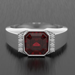 May include: A silver ring featuring a large, square, deep red gemstone. Small, clear gemstones are set either side of the central stone. The ring has a wide band and a modern design.