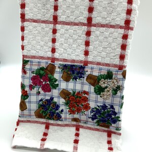 May include: White kitchen towel with red checkered border and a floral print panel featuring potted flowers in shades of red, pink, purple, and green on a blue and white checkered background.