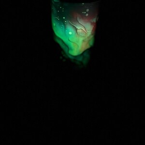 May include: A glowing tumbler with a vibrant design. The upper portion features a blend of green and red hues, with white speckles. The bottom part of the tumbler is a darker shade of green. The background is black.