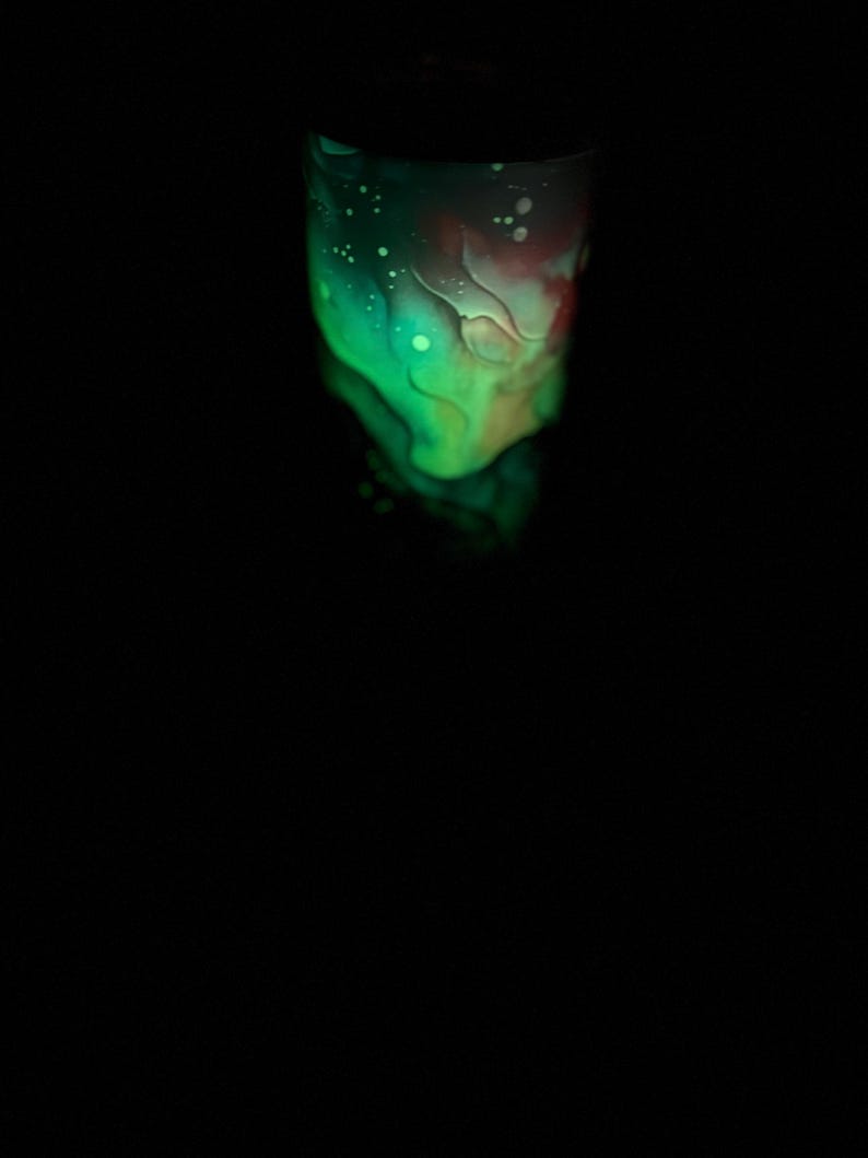 May include: A glowing tumbler with a vibrant design. The upper portion features a blend of green and red hues, with white speckles. The bottom part of the tumbler is a darker shade of green. The background is black.