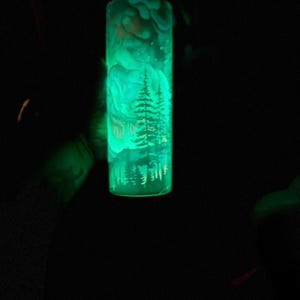 May include: A green, glowing tumbler featuring a forest scene etched into the surface. The design includes trees, mountains, and a house, all illuminated against a dark background. The tumbler appears to be made of glass or a similar material.