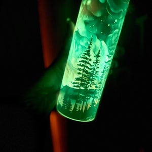 May include: A luminous green tumbler with a forest landscape design, including silhouetted trees and a starry sky. The cylindrical tumbler glows, creating an ethereal effect. The material appears to be glass or a similar transparent substance.