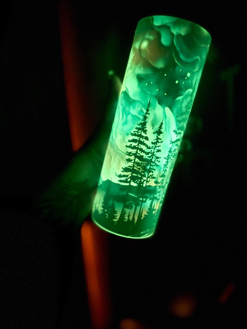 May include: A luminous green tumbler with a forest landscape design, including silhouetted trees and a starry sky. The cylindrical tumbler glows, creating an ethereal effect. The material appears to be glass or a similar transparent substance.